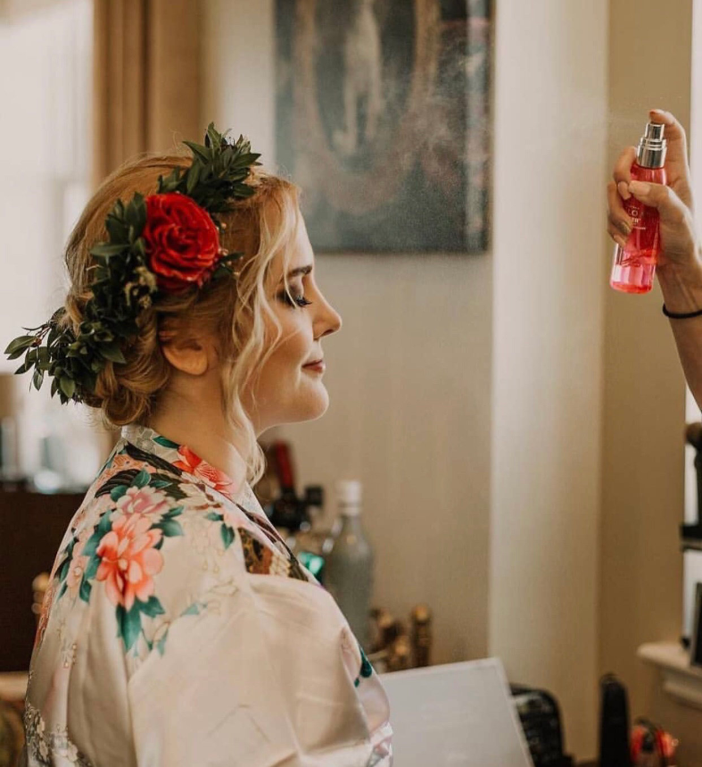 A bride with a floral hair crown sits while her artist is using makeup spray.
