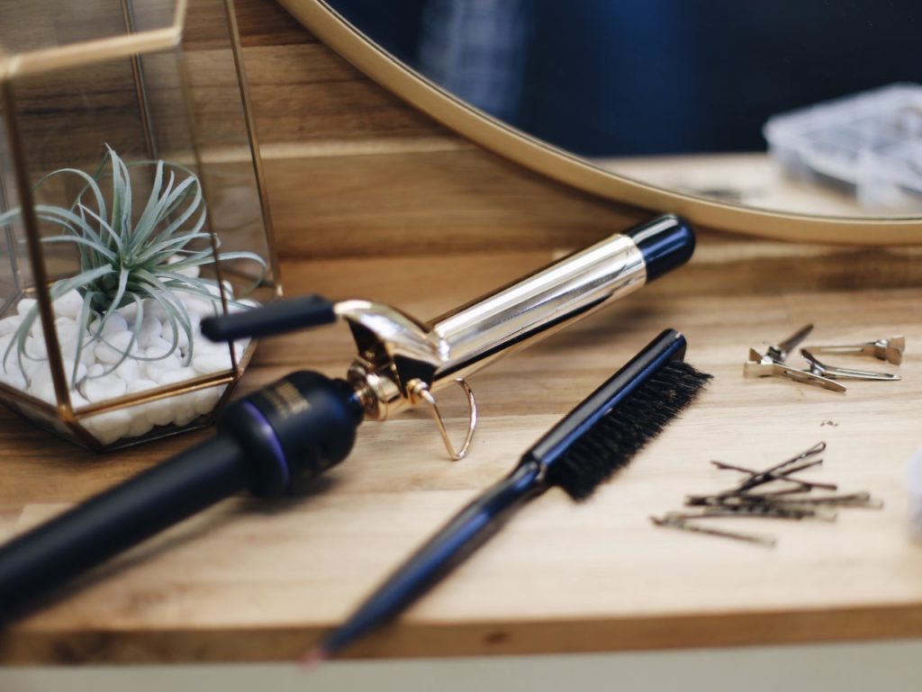 Various hair products (curling wand, bobby pins, and brush) are sitting on a desk with a mirror and plant on it.